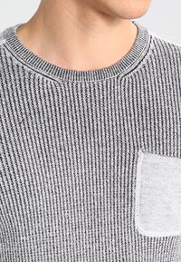 Pull en maille rayé gris et blanc avec un col rond et une poche poitrine fonctionnelle en tissu texturé.