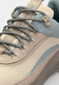 Beige og lyseblå treningssko med teksturert stoff, lagdelt design og unikt snøresystem. Inkluderer gummisåle og polstret krage.