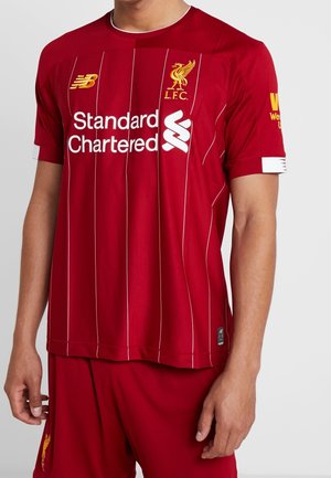 Personne portant le maillot rouge du Liverpool FC avec des fines rayures blanches et des logos de sponsors, assorti à un short rouge.