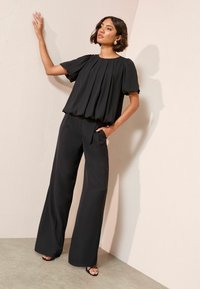 Fekete, pliszírozott, rövid ujjú blúzt és széles lábú nadrágot viselő nő, az egyik kezét a falon támasztva, a másikat a zsebében tartva, szandált viselve.