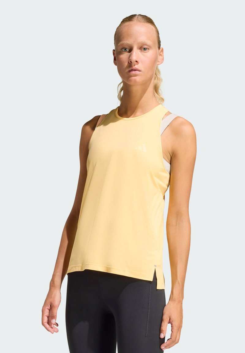 Camiseta deportiva sin mangas amarilla con costuras flatlock, aberturas laterales y logo reflectante en el pecho. Combinada con leggings negros.