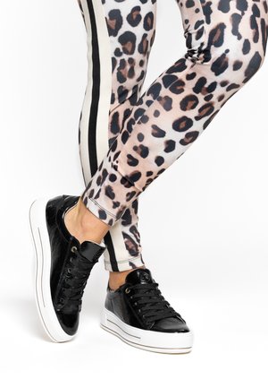 Schwarze Sneakers aus Lackleder mit dicken weißen Sohlen und schwarzen Schnürsenkeln, kombiniert mit Leggings im Leopardenmuster mit seitlichem Streifendetail.