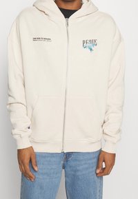 Béžová mikina na zip s kapucí a šňůrkou, s potiskem textu a grafiky na přední straně a předními kapsami. Měkká textilie.