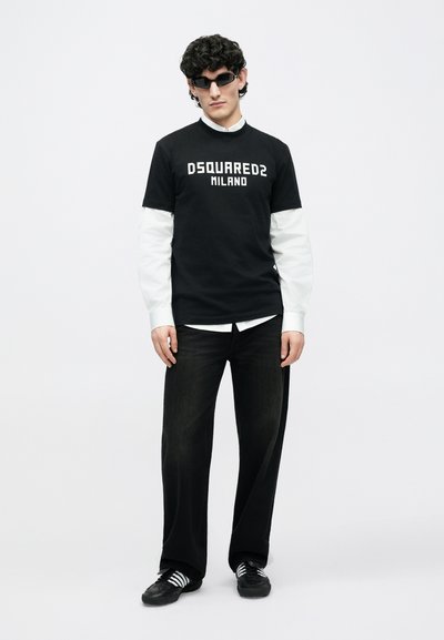 Vīrietis, kas valkā melnus saulesbrilles, melnu DSQUARED2 Milano T-kreklu virs baltas krekla, melnas platām kājām bikses un melnas sporta kurpes ar baltām šņorzēm.