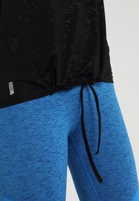 Czarny top z krótkim rękawem z marszczonym dołem i detalem ze sznurkiem, zestawiony z niebieskimi legginsami melanżowymi. Oba zaprojektowane do noszenia podczas aktywności fizycznej.