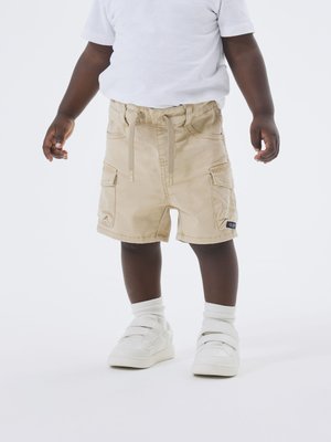 Shorts cargo beige avec taille à cordon, deux poches latérales et détail de label de marque. Portés avec une chemise blanche et des baskets blanches à velcro.