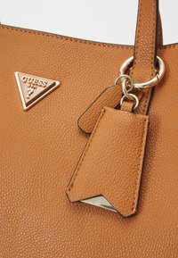 Tan textuur neplederen handtas met een gouden driehoekig logo, voorzien van een afneembare tag en een ronde metalen ring accent.