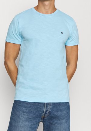 Homme portant un t-shirt à manches courtes bleu clair avec un petit logo rouge, blanc et bleu sur la poitrine, associé à un jean en denim bleu.