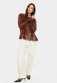 Brown sheer blouse with ruffle detailing, paired with white wide-leg pants and black pointed-toe heels featuring thin straps and accents.