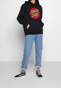 Felpa nera con tasca frontale e cappuccio con cordoncino, caratterizzata da un logo grafico rosso e giallo. Abbinata a jeans azzurri risvoltati e sneakers nere.