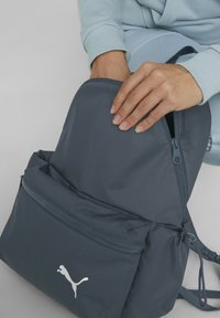 Mochila de nailon azul oscura con un bolsillo frontal con cremallera, que presenta un logo de Puma en blanco. Los tiradores de la cremallera son negros y tiene correas ajustables.