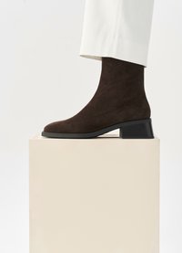 Stivale alla caviglia in suede marrone con punta arrotondata e tacco basso, posizionato su un piedistallo di colore chiaro contro uno sfondo neutro.
