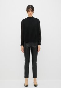 Top nero a maniche lunghe con pieghe verticali, collo alto; abbinato a pantaloni in pelle nera aderenti e scarpe nere con punta e dettagli dorati.
