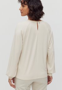 Beige langermet top med et nøglehul i ryggen, blød tekstur og løs pasform. Har rund halsudskæring og manchetærmer.