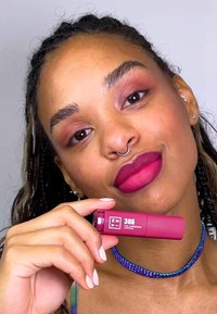 Langvarig leppestift i et sylindrisk rosa rør, holdt av en person. Fargen ser dristig og matt ut på leppene. Minimalistisk design med klar merking.