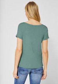 Grön t-shirt med kort ärm och rundad halsringning samt viksömmar, tillverkad av ett mjukt material, i kombination med blå jeans.