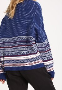 Niebieski sweter dzianinowy z białymi wzorami geometrycznymi oraz paskami w kolorach czerwonym, białym i niebieskim. Luźny krój z prążkowanymi mankietami i dołem.