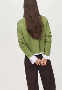 Pull vert court à manches longues superposé sur une chemise blanche, associé à un pantalon marron foncé, vu de dos. Cheveux longs et ondulés visibles.