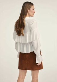 Biała bluzka z długim rękawem i warstwowymi falbanami, zestawiona z brązową spódnicą z zamszu. Bluzka posiada kołnierzyk i rozkloszowane rękawy.