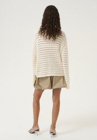 Stickad tröja i krämfärg med en öppen vävdesign, långa ärmar och scallopkant, parat med beiga shorts och minimalistiska sandaler.
