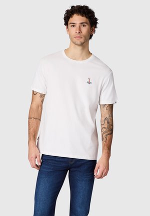 Man draagt een wit T-shirt met een kleine kleurrijke afbeelding op de borst en donkerblauwe spijkerbroek, staand tegen een effen lichte achtergrond.