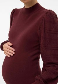 Burgundy stickad graviditetströja med långa ärmar; texturerade puffärmar har ett mönstrat design, hög hals och figurnära silhuett.