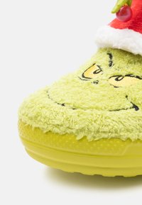 Green slipper featuring a textured, fluffy upper with a Grinch face design. It has a yellow sole and a red Santa hat accent on top.
