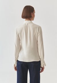 Long-sleeve blouse in cream silk with a collar, pleated shoulders, and buttoned cuffs, paired with dark denim trousers.