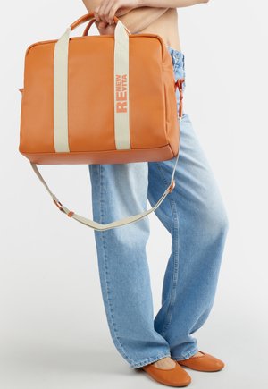 Persona con jeans azzurri chiari e ballerine arancioni che tiene una grande borsa arancione con cinghie beige e la scritta "RENEW EVITA".