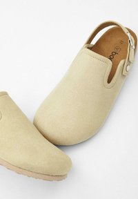 Sabots en suede beige avec un dos ouvert, une conception à bout arrondi et une sangle à boucle en accent. Caractéristiques d'une semelle extérieure texturée pour une bonne adhérence.