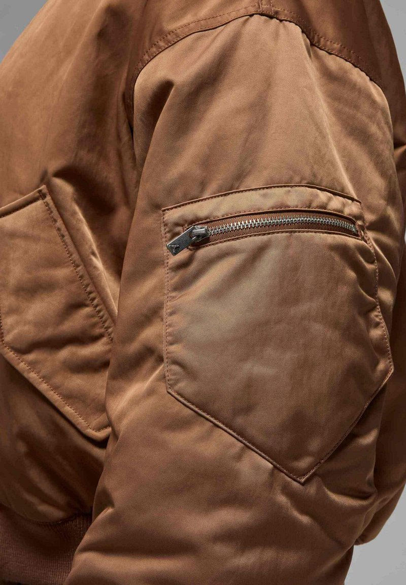 Veste bombardier marron en tissu texturé, avec des coutures d'épaule inclinées et une poche zippée sur le bras pour un détail supplémentaire.