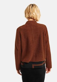 Brun, ludig texturerad jacka med avslappnad passform, som har en krage och en mönstrad svart midja med orange ränder.