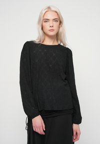 Γυναίκα με μακριά ξανθά μαλλιά, που φοράει μια μαύρη υφασμάτινη μπλούζα με μακριά μανίκια και μαύρη φούστα, στέκεται απέναντι σε ένα απλό ανοιχτό φόντο.