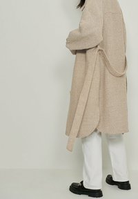 Beige geweven jas met een gestructureerde afwerking, voorzien van een strikceintuur, zijzakken en een gebogen zoom, gecombineerd met witte broeken en zwarte schoenen.