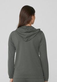 Mujer con cabello largo castaño que lleva una sudadera con capucha de color verde oscuro, vista desde atrás sobre un fondo claro y liso.