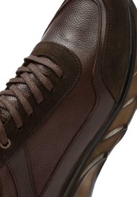 Zapatilla de cuero marrón con acabado texturizado, que presenta acentos de ante marrón oscuro texturizado, cordones redondos y una suela de goma curvada.