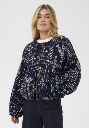 Blondynka ubrana w granatową kurtkę zapinaną na zamek z białymi wzorami paisley i kwiatowymi, w zestawieniu z ciemnoniebieskimi dżinsami.