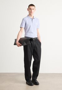 Hellblaues Poloshirt mit weißen Akzenten, schwarze Hose und schwarze Lederclutch. Das Model steht auf einer glatten grauen Oberfläche vor einem schlichten Hintergrund.