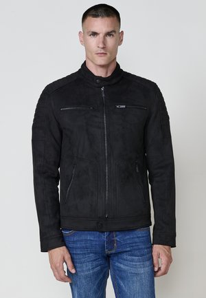 Chaqueta de ante negra con cuello alzado, cierre frontal con cremallera y dos bolsillos con cremallera. Presenta hombros texturizados y un diseño ajustado.