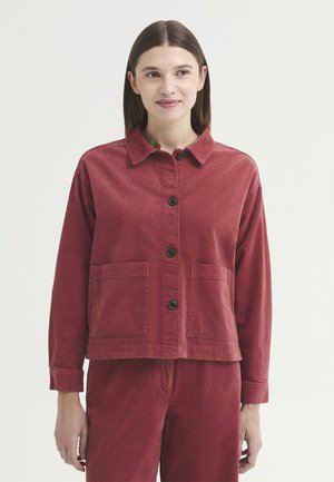 Femme portant une veste boutonnée en velours côtelé rouge avec deux poches à l'avant et un pantalon rouge assorti, debout devant un fond uni.