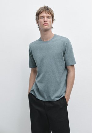 Junger Mann mit kurz zerzaustem Haar, der ein hellgraues kurzärmeliges Hemd und schwarze Hosen trägt, die Hände in die Taschen gesteckt, vor einem schlichten hellen Hintergrund.