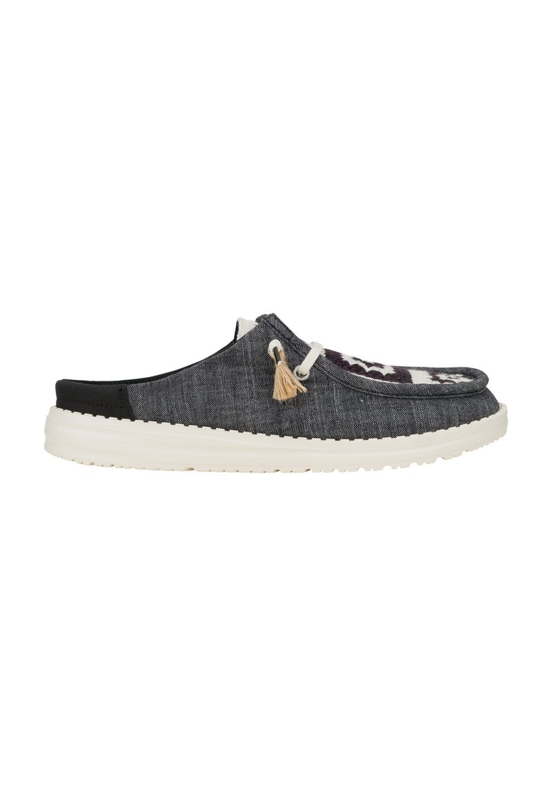 Zapato tipo slip-on de tela gris con parte trasera negra y suela de goma blanca. Presenta borlas decorativas y un diseño frontal con patrones.