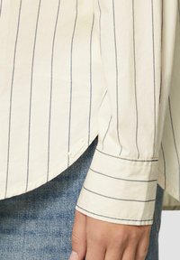 Gestreepte lange mouwen shirt in lichtroom met donkerblauwe verticale strepen. Heeft een ronde zoom en knoopmanchetten, gemaakt van lichte stof.