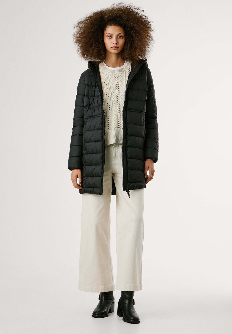 Cappotto bombato nero con cappuccio, caratterizzato da sezioni trapuntate, cerniera frontale e maniche lunghe, indossato sopra un maglione di maglia color crema e pantaloni larghi color crema.