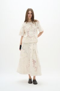 Weißes, floral gemustertes Oberteil und dazu passender langer Rock aus strukturiertem Stoff. Das Model trägt dunkle Slipper und hält eine schwarze Clutch.