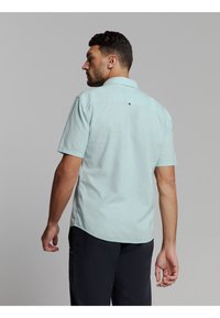 Man met de rug naar de camera, in een lichtblauwe korte mouwen overhemden en donkere broek, tegen een effen grijze achtergrond.