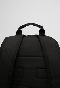 Zaino in tessuto nero con schienale imbottito, spallacci regolabili e manico superiore. Il materiale sembra resistente, con una superficie liscia e cuciture rinforzate.