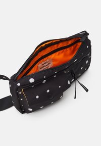 Banane noire à pois blancs, intérieur orange, compartiment principal zippé, poche avant avec bouton-pression et sangle noire.