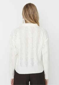 Weißer Strickpullover mit Zopfmuster, verziert mit Lochstrickdetails und einem Kragenhals. Bündchen und Saum sind gerippt und runden das Design ab.