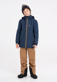 Veste imperméable bleue avec des accents beige, dotée d'une fermeture éclair à l'avant et de poches. Pantalons de ski beige avec des sections inférieures de jambe brun foncé et des bottes noires.